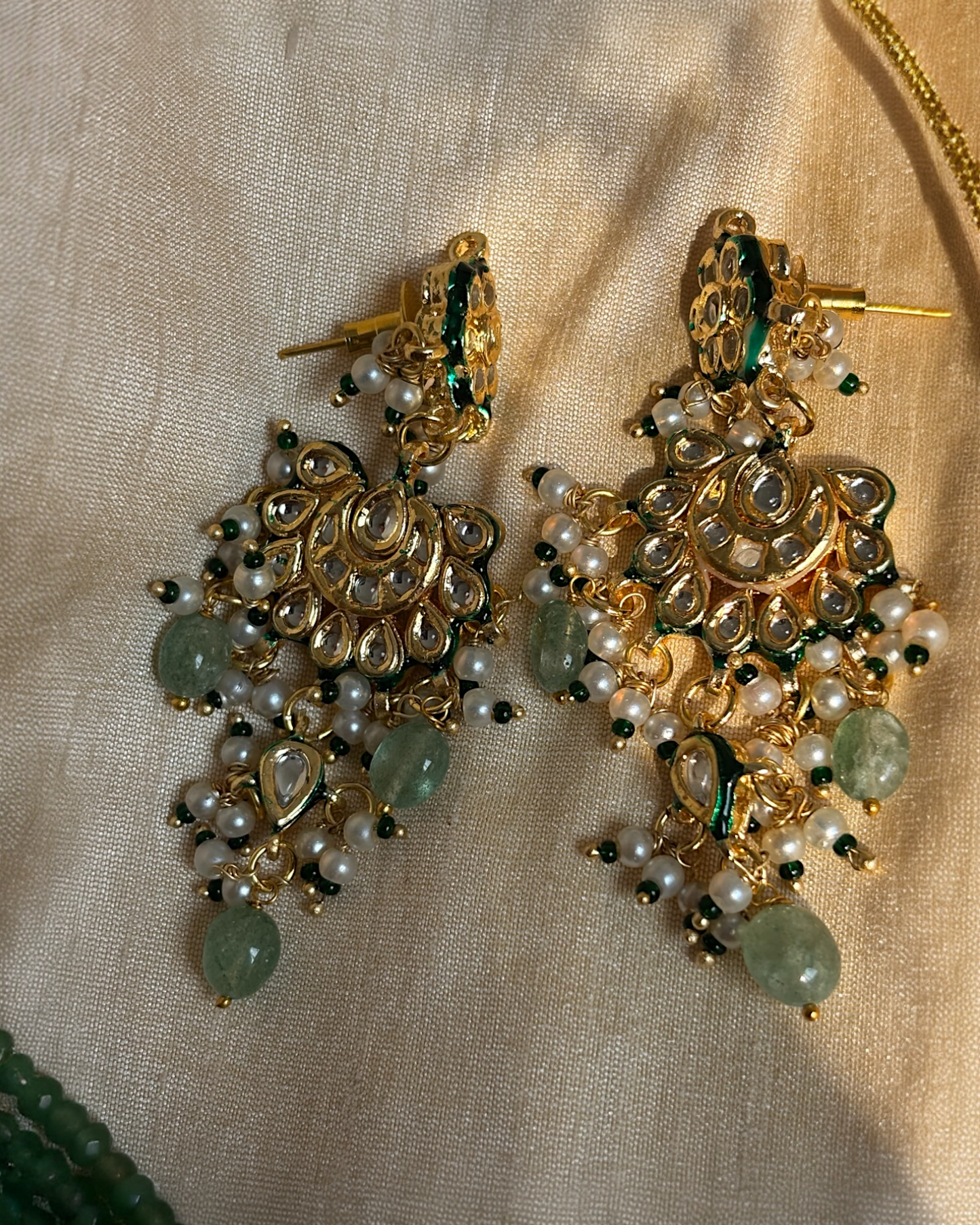 Ornate gold earrings with pearls and green stones on a beige fabric background