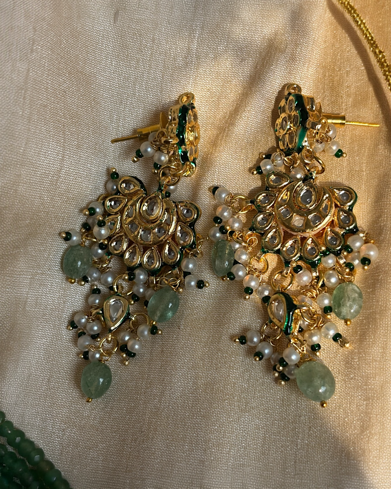 Ornate gold earrings with pearls and green stones on a beige fabric background